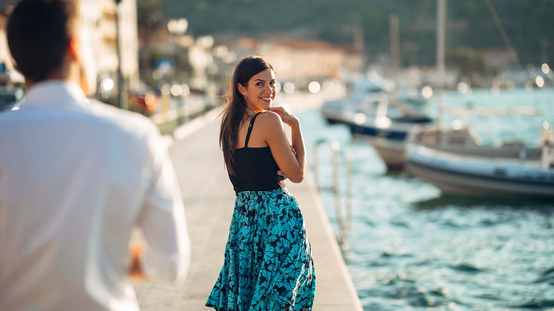 Brunette woman in blue skirt and black shirt looking back at a man in white shirt and smiling. They are at the docks.